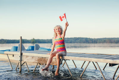 Girl sitting on dock pier by lake and waving canadian flag. canada day holiday on july outdoor.