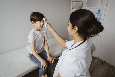 Female pediatrician measuring temperature of boy through infrared thermometer at hospital