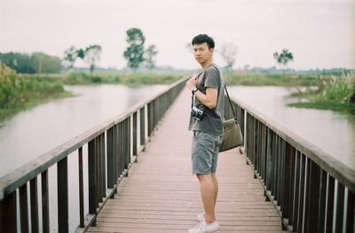 Portrait of young man standing on footbridge amidst lake