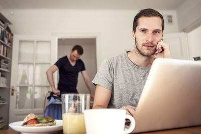 Portrait of gay man sitting with laptop at breakfast table with partner ironing in background