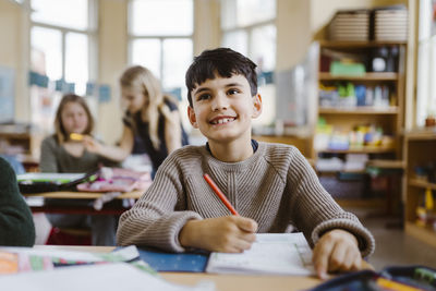 Smiling male pupil holding pencil while sitting at desk in classroom