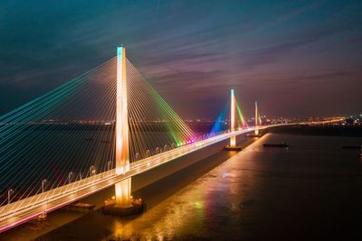 Illuminated suspension bridge over river against sky at night