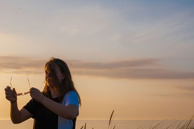 Smiling woman holding sunglasses standing against sky