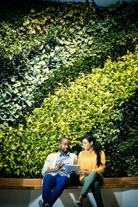 Businessman and woman sitting in front of green plant wall, using digital tablet