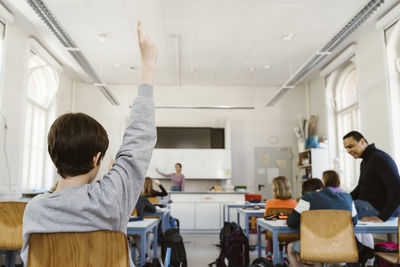 Rear view of schoolboy raising hand while attending lecture in classroom