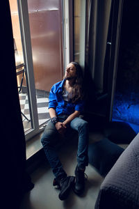 Man with long hair and beard sits at home on a chair by the window in a blue shirt and jeans