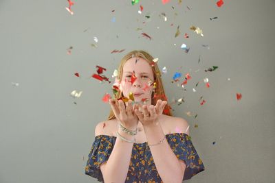 Portrait of young woman blowing confetti against wall