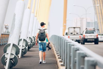 Rear view of young man holding camera while walking on bridge against sky