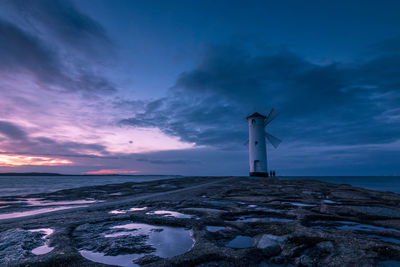 Traditional windmill by sea against cloudy sky during sunset
