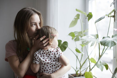 Mother kissing daughter while looking through window at home