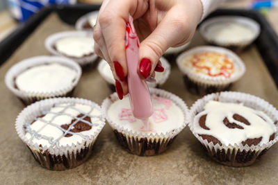 A woman squeezes colored frosting from a tube onto chocolate brown cupcakes covered white frosting.