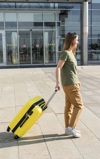 Young woman traveler carrying a yellow suitcase next to the entrance to the airport outside luggage