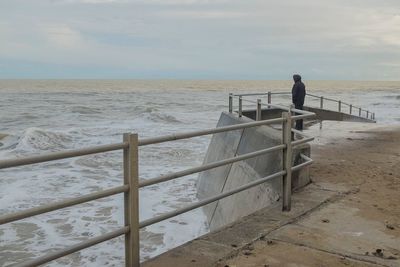 Man standing on railing by sea against sky