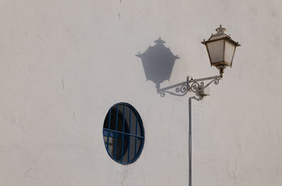 Low angle view of street lamp and shadow with small round blue window on white wall