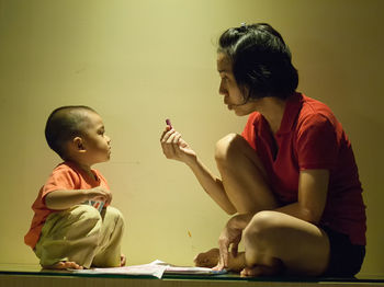 Mother showing crayon to son while sitting on table by wall