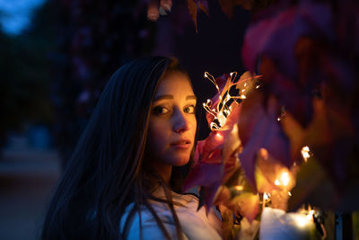Portrait of young woman with illuminated string lights by plants at night