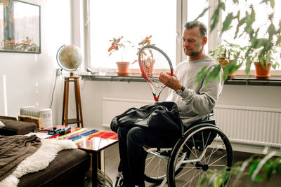 Thoughtful mature man holding tennis racket while sitting on wheelchair at home