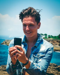 Smiling man using smart phone at beach against sky