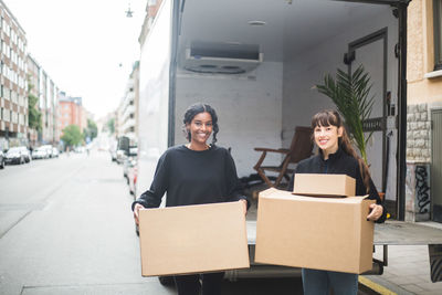 Portrait of smiling female movers unloading boxes from truck on street in city