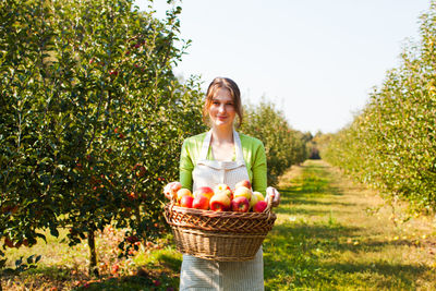 Smiling young woman holding apple in basket against trees