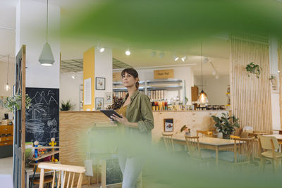Businesswoman holding digital tablet looking away while standing in cafe