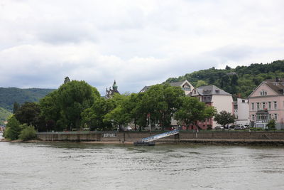 Trees and buildings by river against cloudy sky