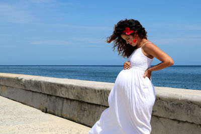 Pregnant woman standing caressing her pregnant belly by sea against sky