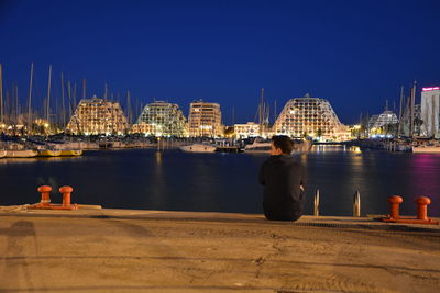 Rear view of man sitting on pier at harbor against clear blue sky at dusk