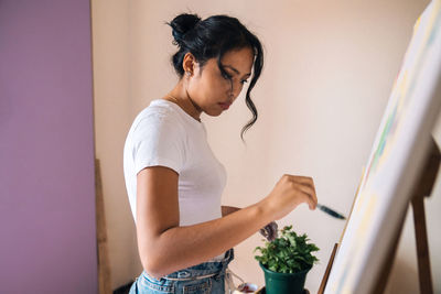 Side view ethnic female in casual clothes painting on canvas with multicolored paints and brush in light small room