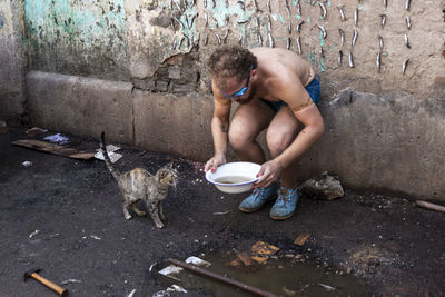 Full length of a shirtless man giving water to a cat against a wall