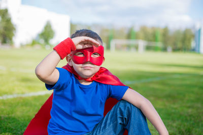 Portrait of boy in costume and mask shielding eyes while sitting on field