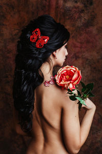 Rear view of shirtless young woman holding flower standing against wall