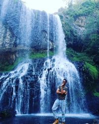 Portrait of smiling young man standing on rock against waterfall