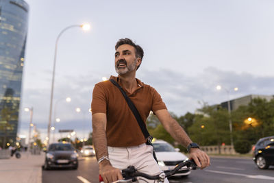 Mature businessman with bicycle standing at roadside in city on sunset