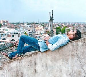 Side view of man lying on retaining wall in city against sky