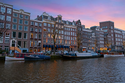 Boats moored in river by buildings against sky during sunset