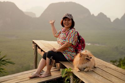 Full length of woman sitting on wood against mountains with dog. 