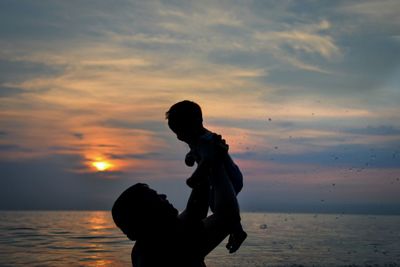 Silhouette man with toddler son at beach against sky during sunset