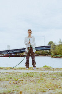 Full length portrait of confident young non-binary person holding garden hose near river