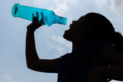 Silhouette of a girl drinking last drop of water from a empty blue plastic bottle under sky