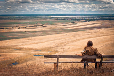 Rear view of person sitting on a bench overlooking field against cloudy sky