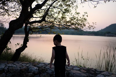 Rear view of boy standing at lakeshore during sunset