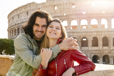 Young couple traveling to rome. the couple smiles and hugs for a selfie in front of the colosseum.