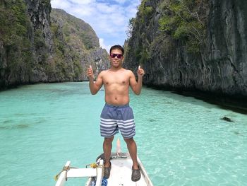 Portrait of smiling shirtless man showing thumbs up while standing on ship bow on sea