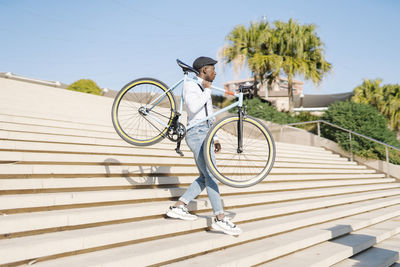 Young man carrying bicycle and moving down staircase on sunny day