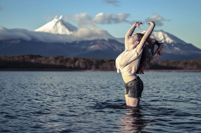 Side view of young woman with arms raised standing in lake against snowcapped mountain