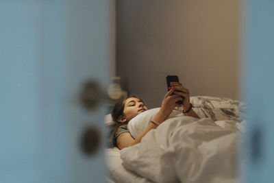 Girl using smart phone while lying on bed in bedroom at modern home