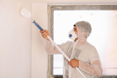 Man in protective workwear and mask working with paint roller standing against white wall at home