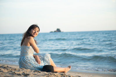 Portrait of young woman sitting at shore of beach against sky