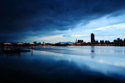 Illuminated buildings by sea against sky at dusk